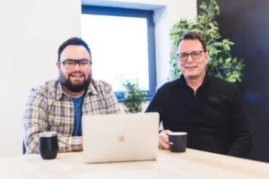 Shaun and Callum having a meeting with their laptops sat at a wooden table, while smiling at the camera