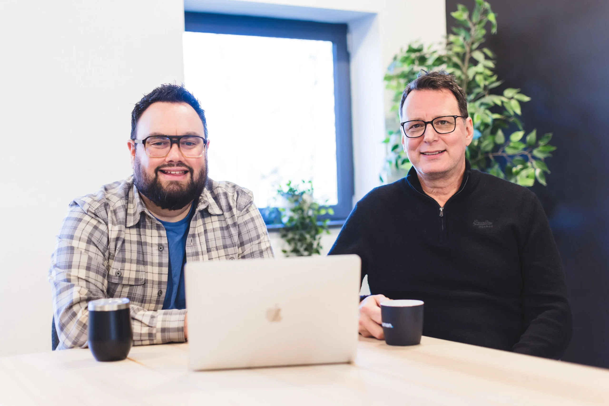 Shaun and Callum having a meeting with their laptops sat at a wooden table, while smiling at the camera