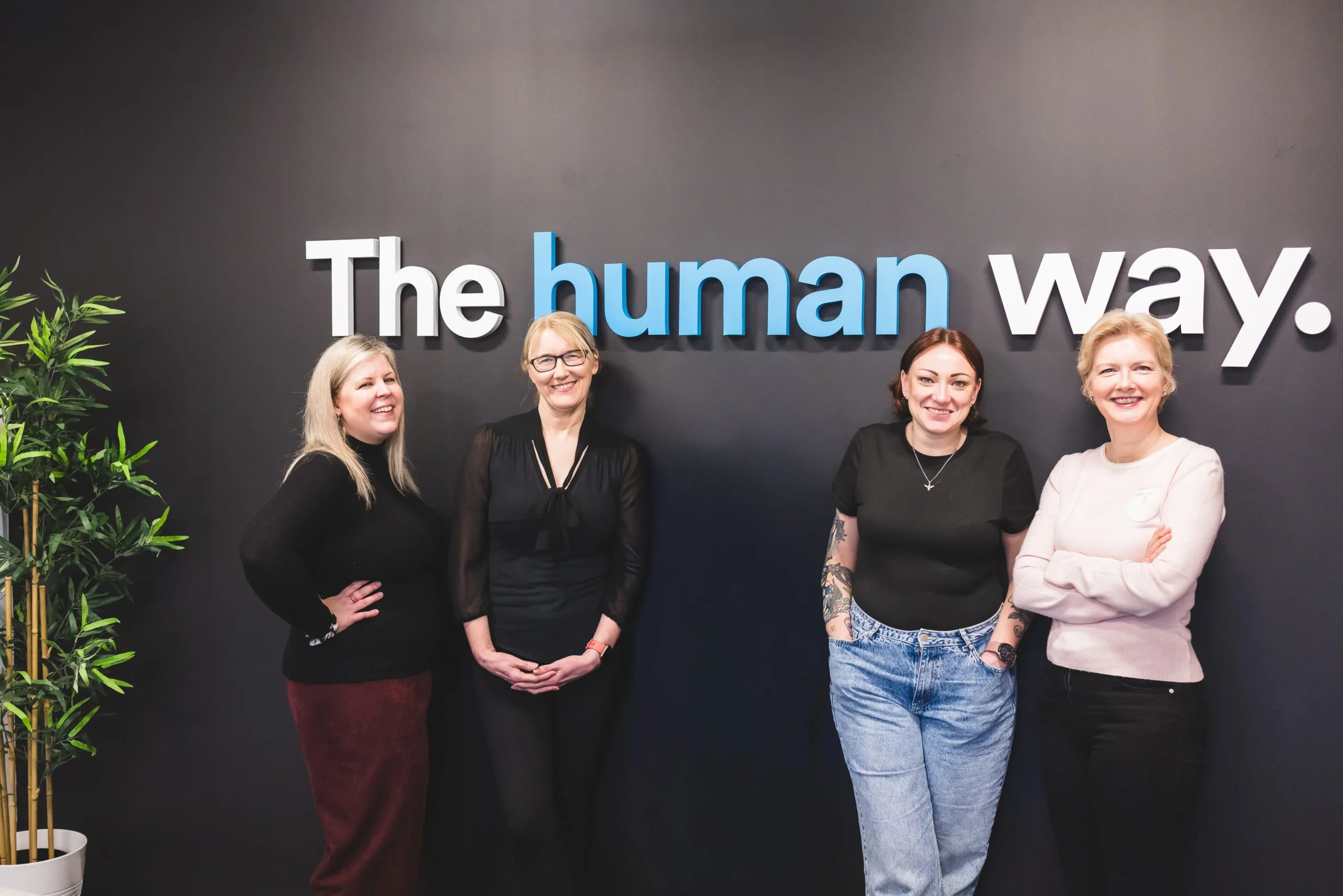 Four female members of Damteq's management team stood on the office boardroom, behind them is a sign with Damteq's slogan that says 'the human way'