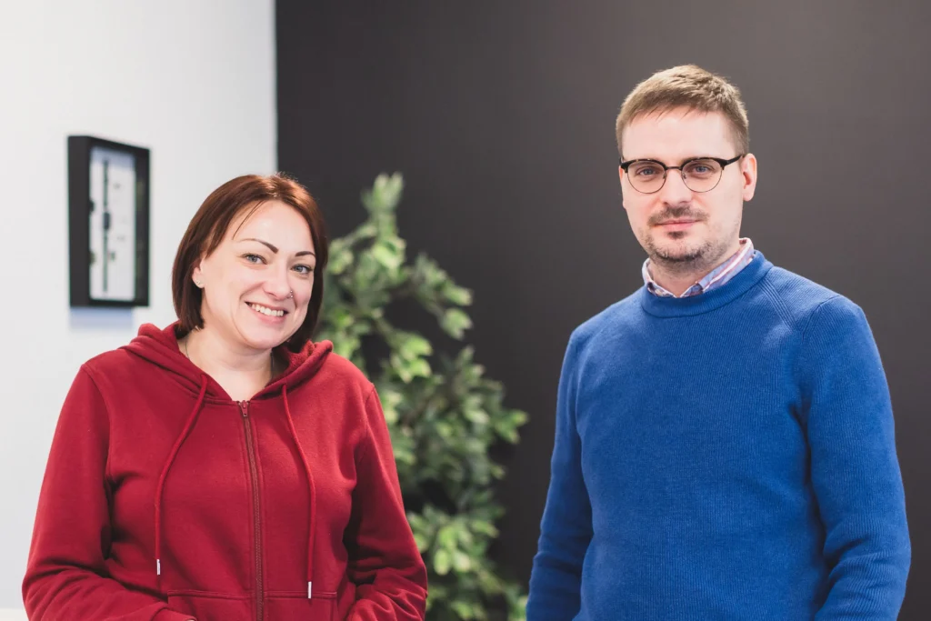 Closeup of Gemma and Milan stood in an office smiling at the camera