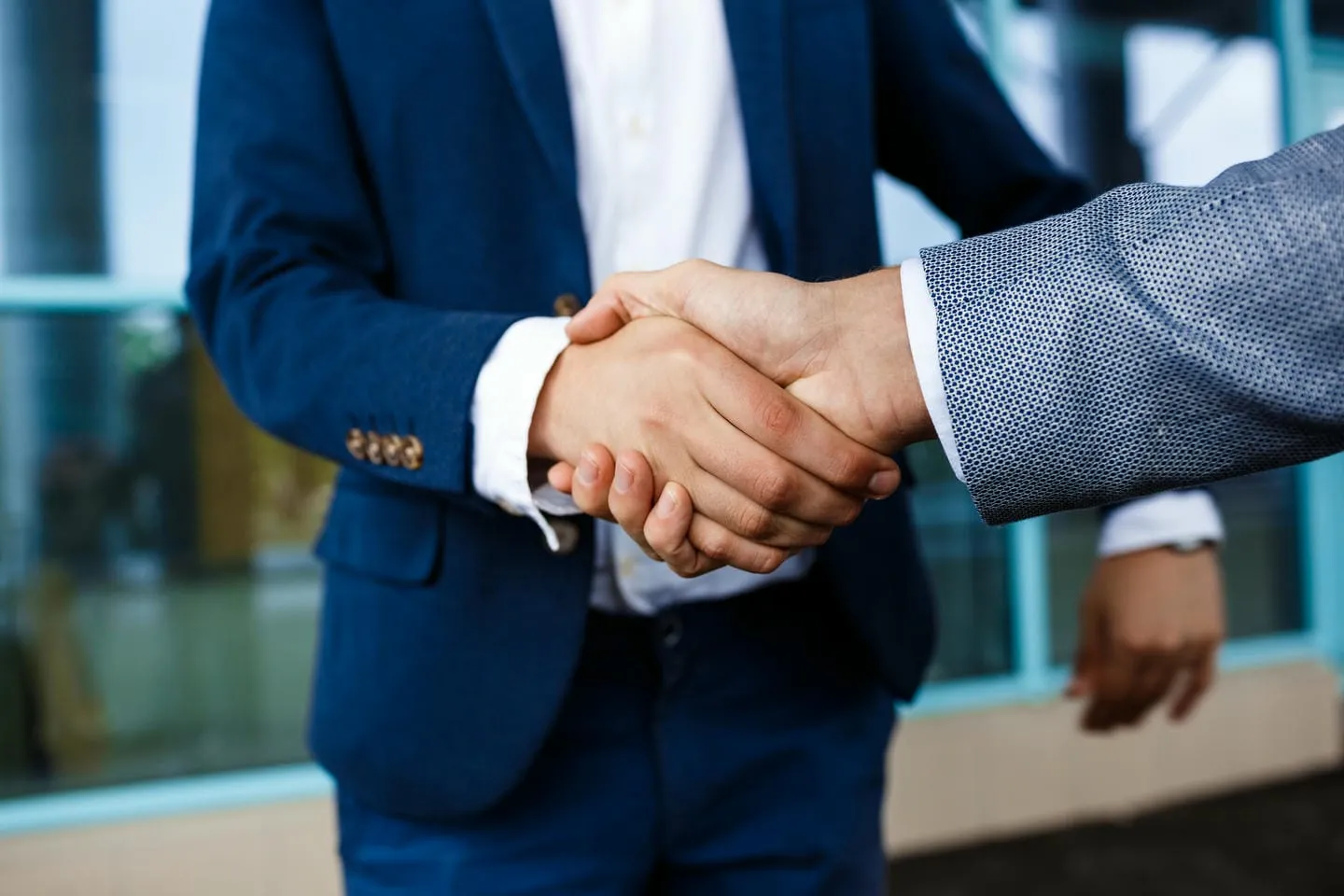Two businessmen in blue suits, shaking hands outside of a modern office building